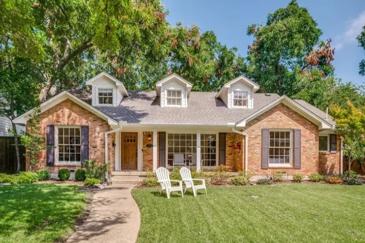 Single-story brick home with a front porch, tall trees and two chairs on the lawn